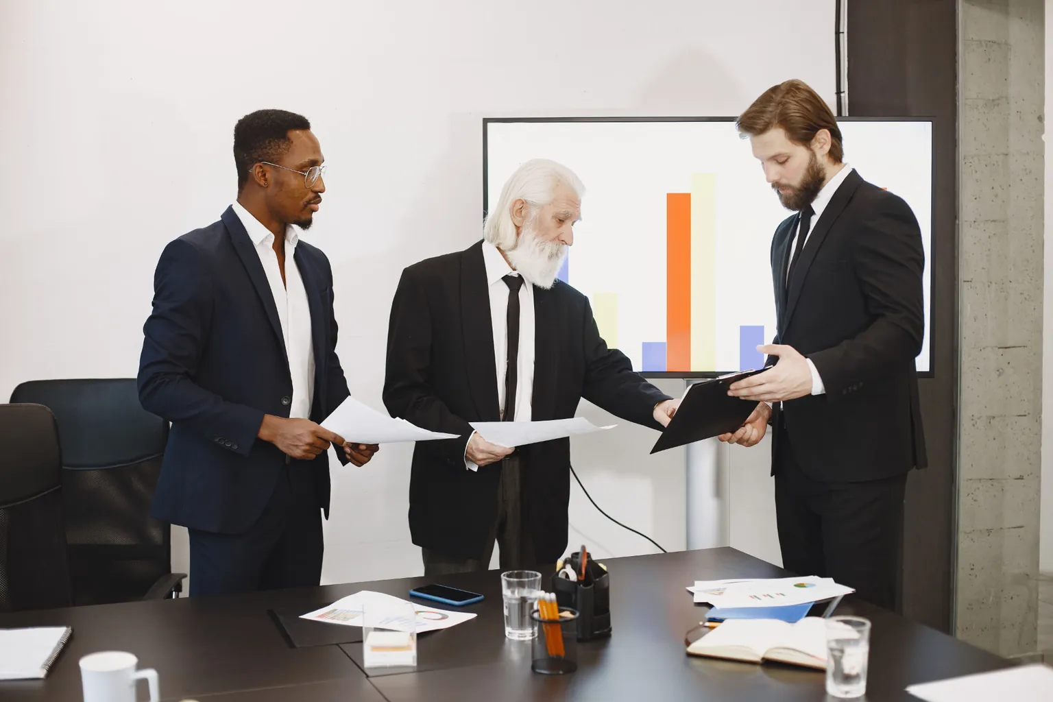 Three business professionals reviewing sales documents together in a modern office with a bar chart on a screen behind them