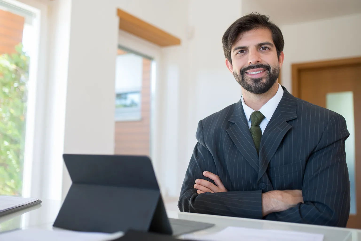 Smiling sales executive with arms crossed seated at a desk with a tablet