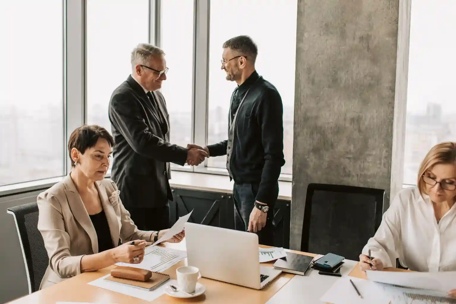 Two consultants shaking hands across a conference table while colleagues review documents